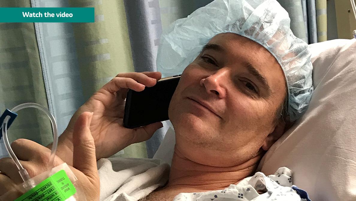 Chet Kitchen on a cell phone, wearing a hospital gown and hair covering.