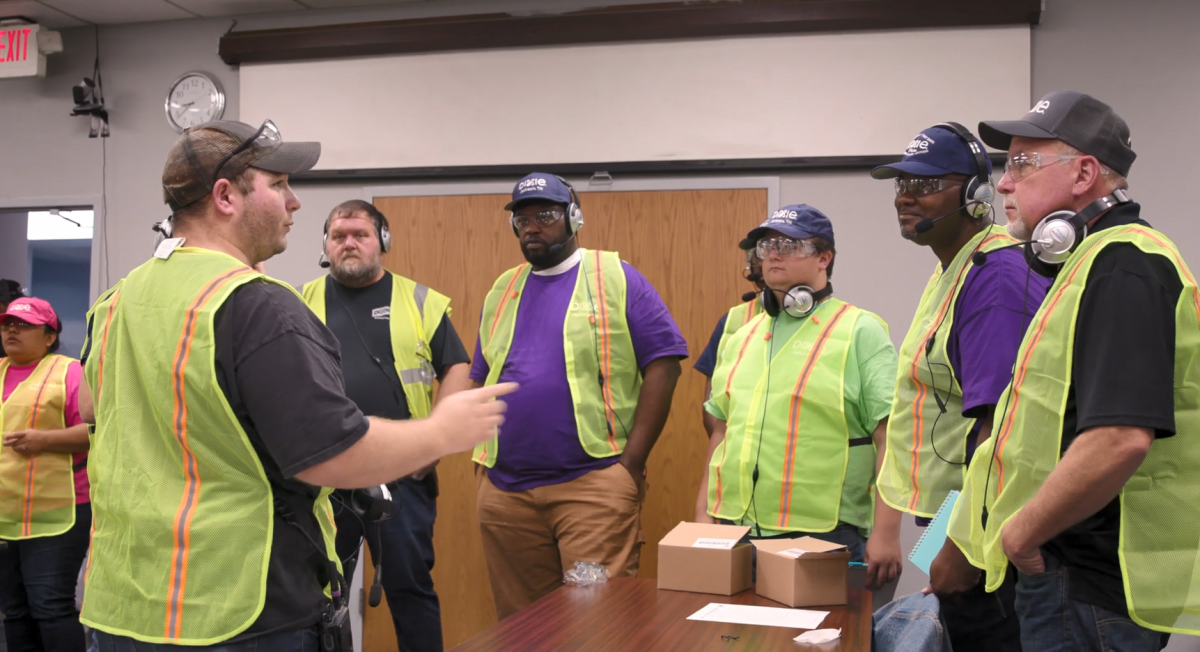 A group of people in high-vis vests gathered in a room, listening to one speaking.