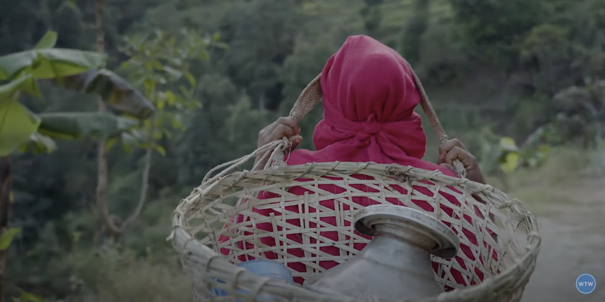 person with a pink head covering carrying a water jug in a wicker basket