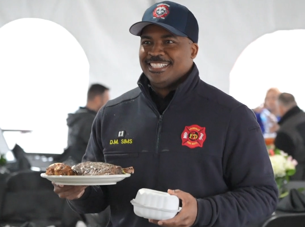 A smiling person in firefighter jacket holding a plate and container of food.