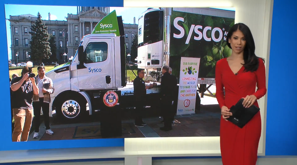 A reporter, on a screen behind them people meeting around a semi-truck outside with Sysco on the side.