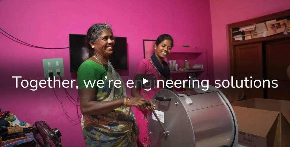 Two people with a washing machine in front of a bright pink wall