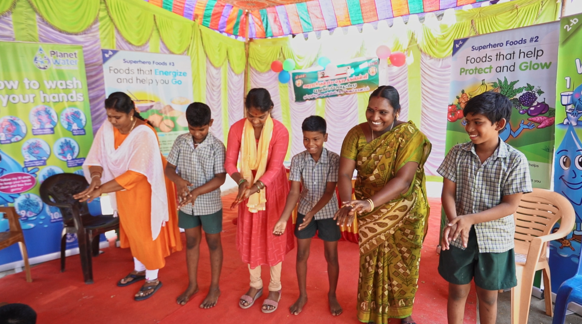 A group of smiling adults and kids demonstrating hand washing.  Colorful signs behind them. 