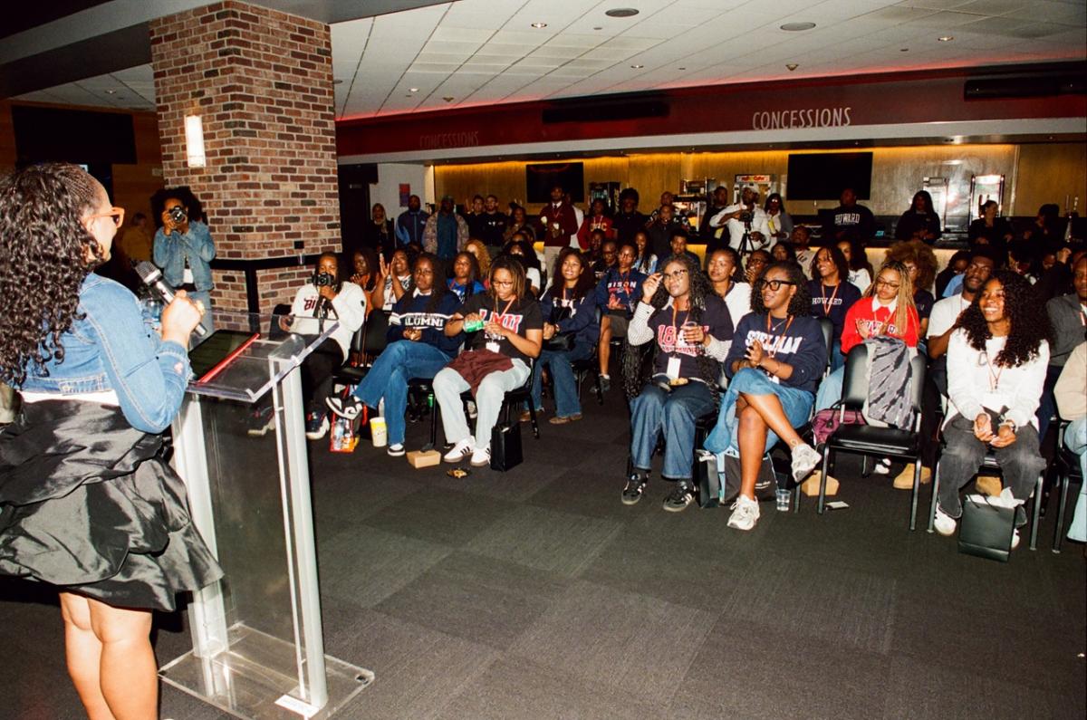 Person speaking at a podium inside an arena space while a seated audience of students listens during a program or presentation.