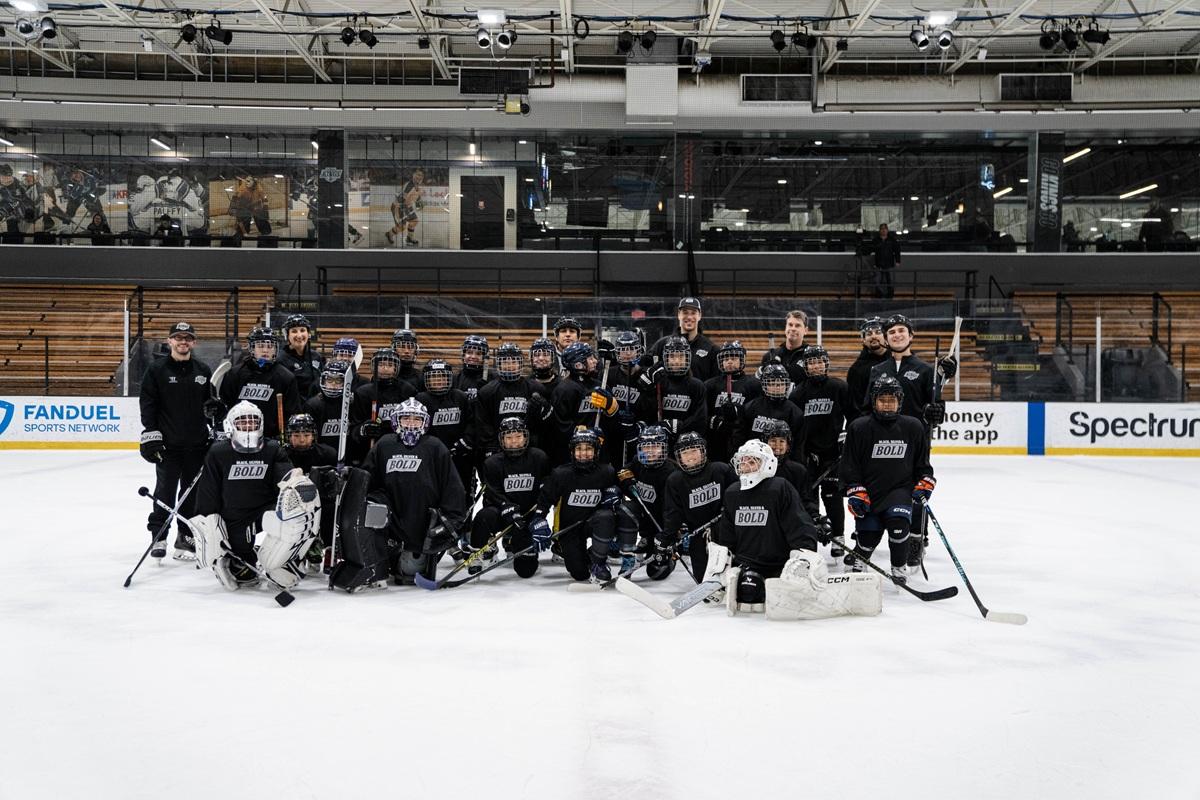 Group photo of youth participants and coaches in Black, Silver & Bold jerseys posing together on the ice rink.