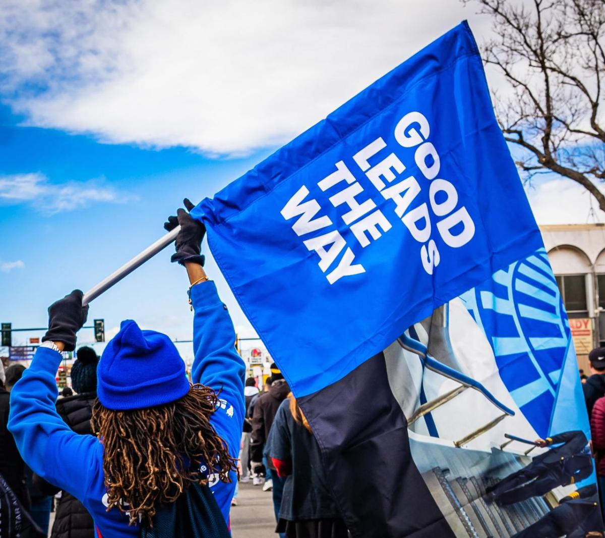 Person wearing blue holds a blue flag with the words, "good leads the way" printed in white on the flag
