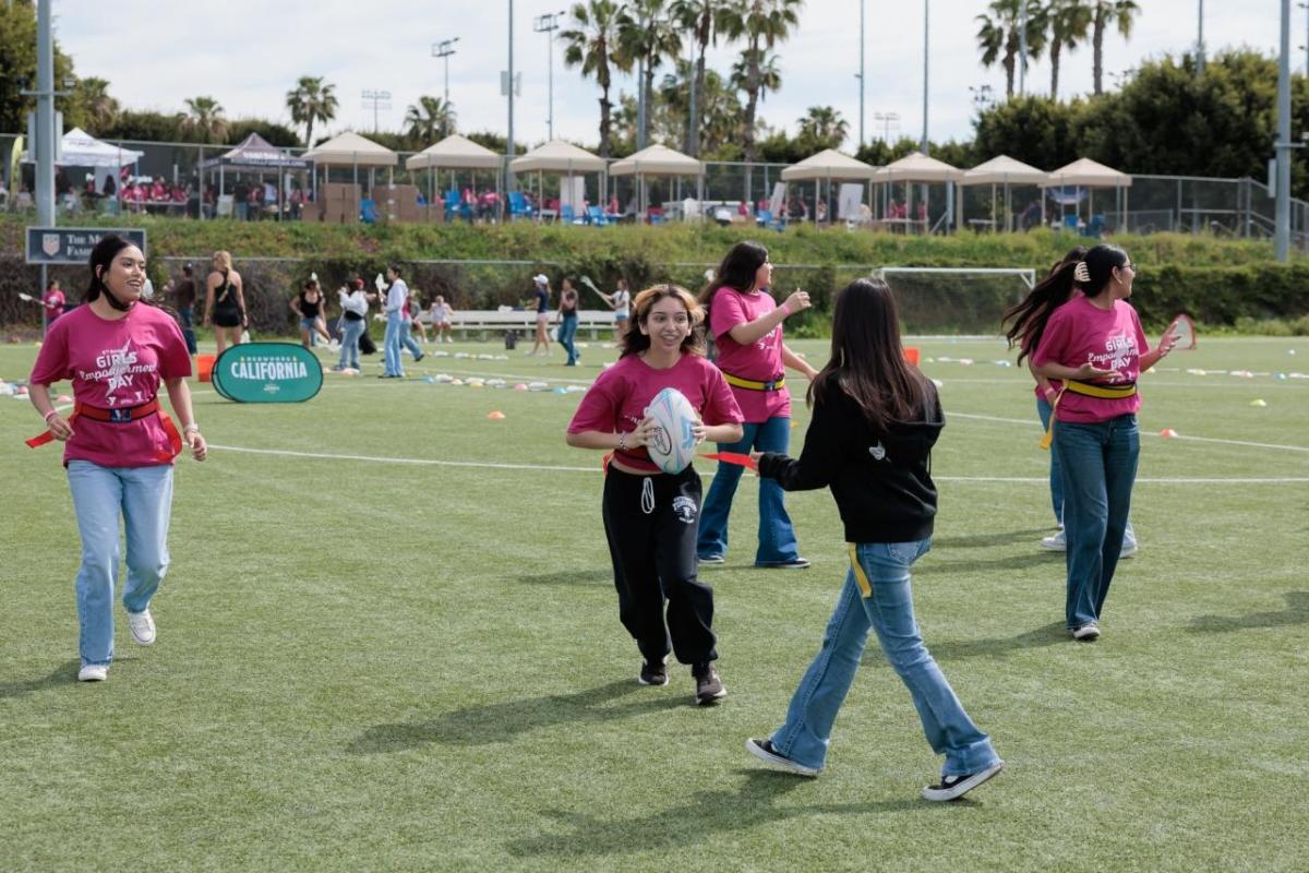 Girls wearing pink “Girls Empowerment Day” shirts play flag football on a soccer field during a youth sports clinic.