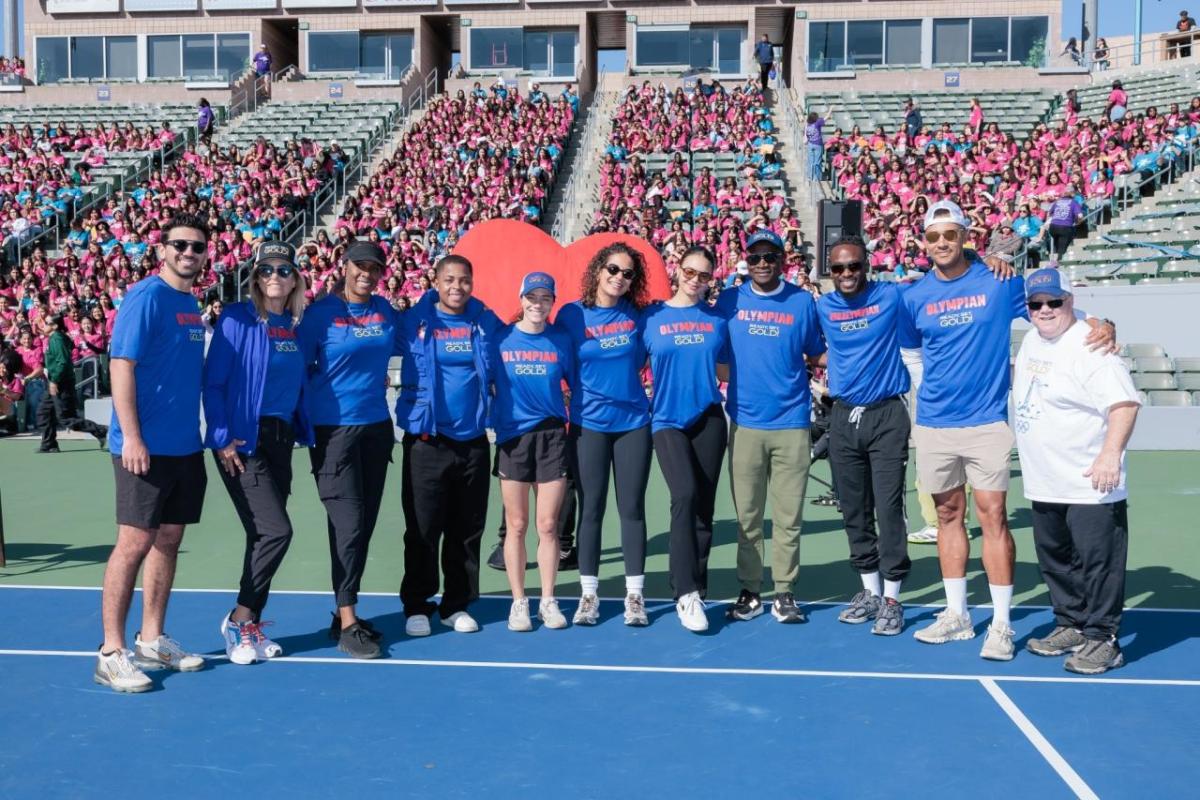 Olympians and Paralympians wearing blue “Olympian Ready, Set, Gold!” shirts pose for a group photo on the field in front of a stadium crowd of girls attending Girls Empowerment Day.
