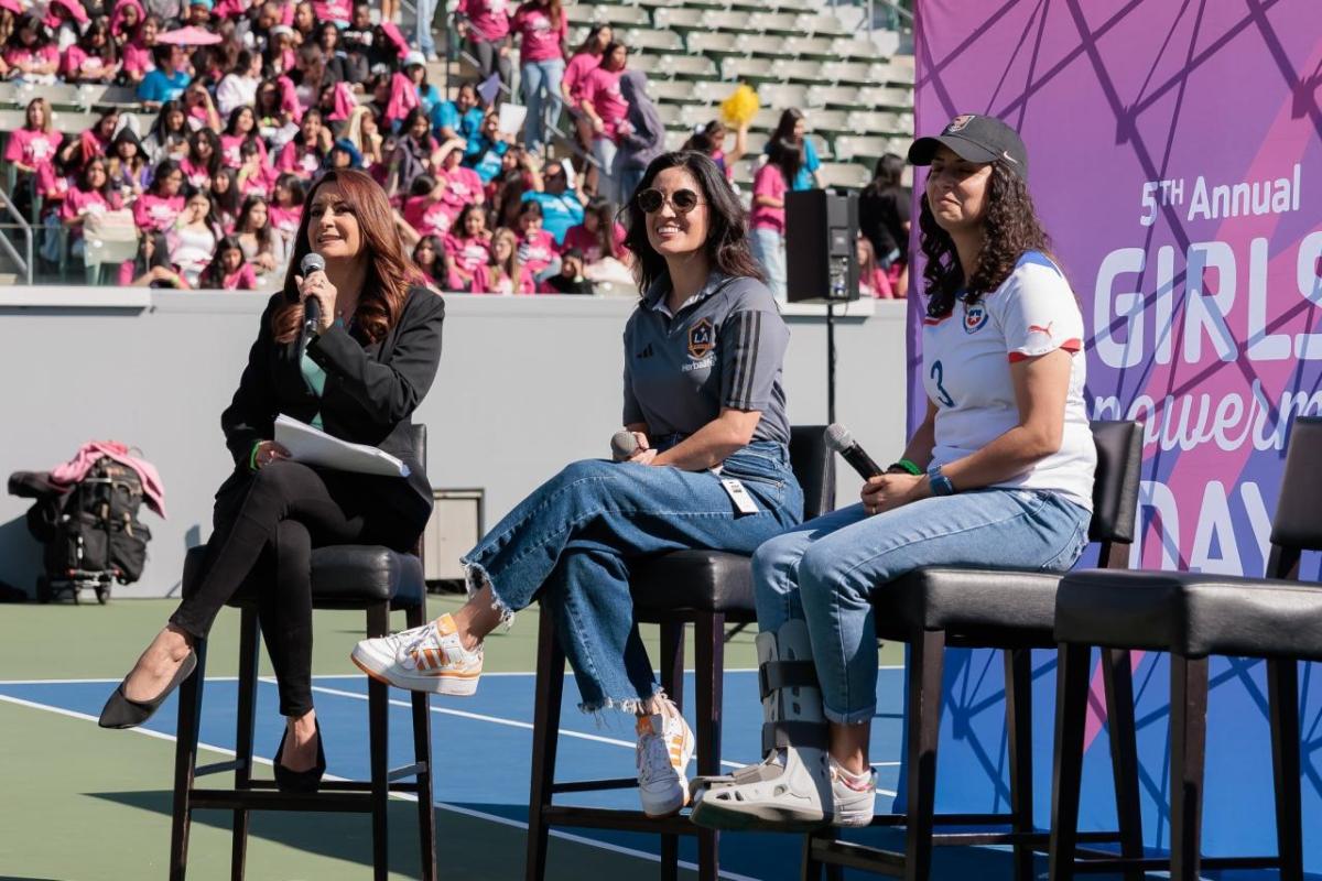 Three women participate in a panel discussion during the 5th Annual Girls Empowerment Day while a crowd of attendees sits in the stadium stands behind them.