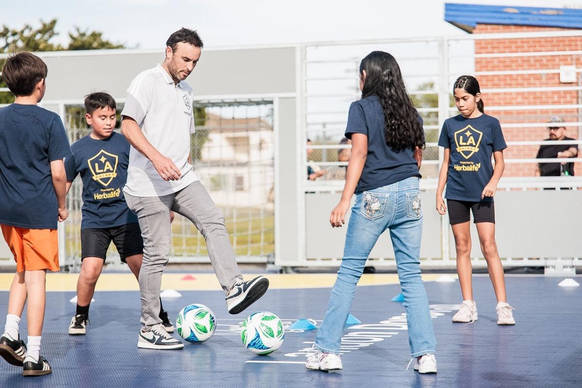 Landon Donovan coach demonstrates ball control while children in team shirts practice with multiple soccer balls on the court.