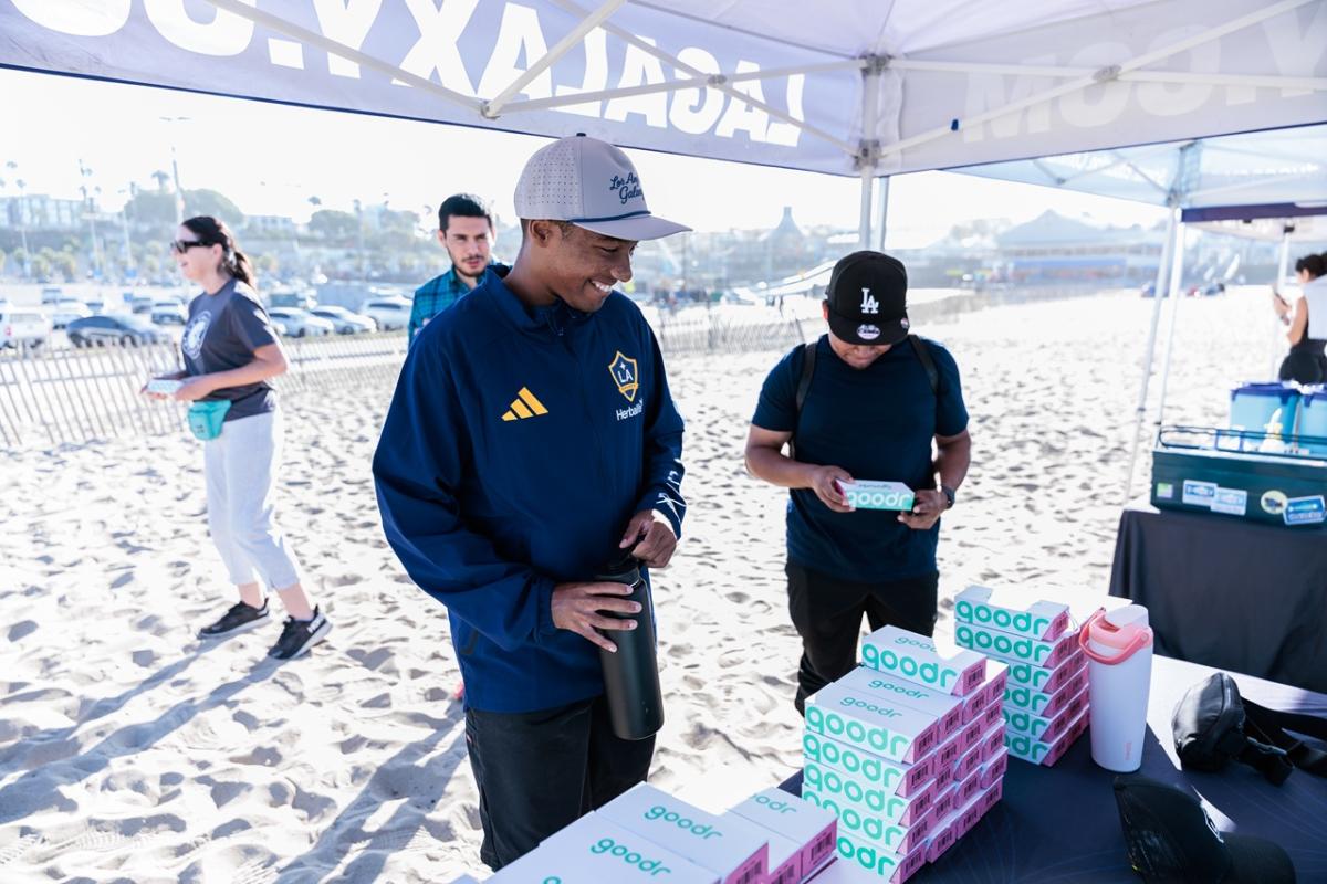Volunteers stand under an LA Galaxy canopy on the beach, with one person receiving items from a table stacked with Goodr sunglasses boxes.