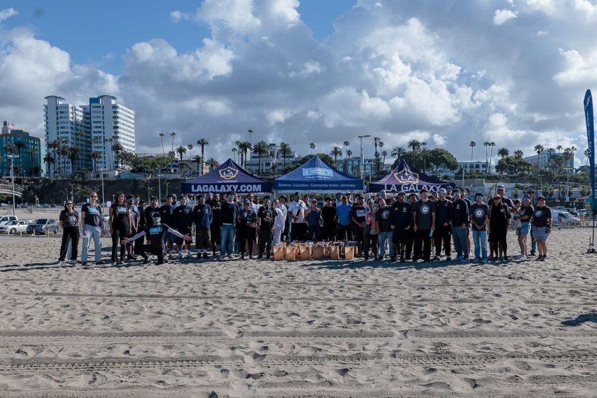 A large group of volunteers pose on the beach behind several trash buckets, with LA Galaxy tents and palm trees visible in the background.