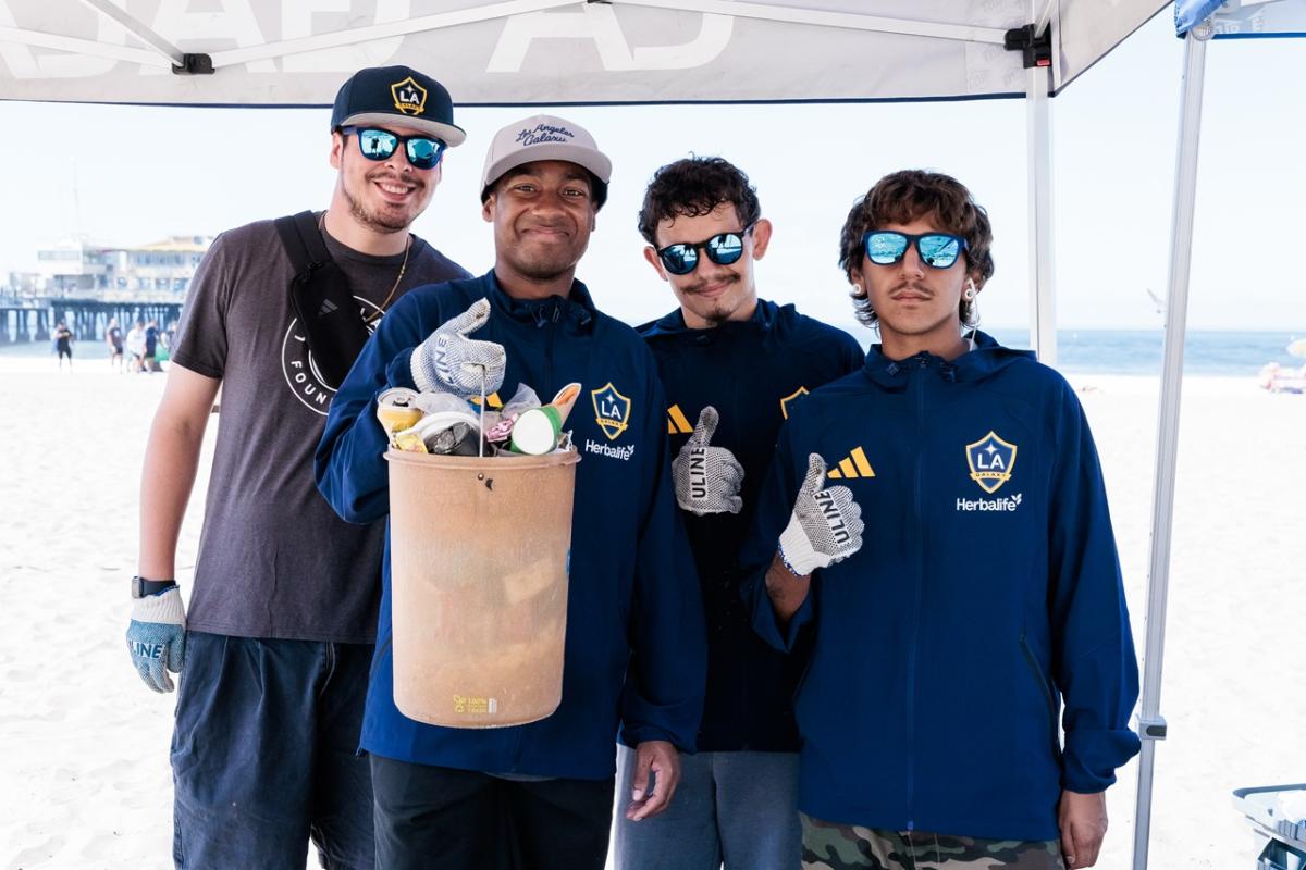 Four volunteers wear LA Galaxy jackets and gloves while holding a bucket filled with collected trash during a beach cleanup.