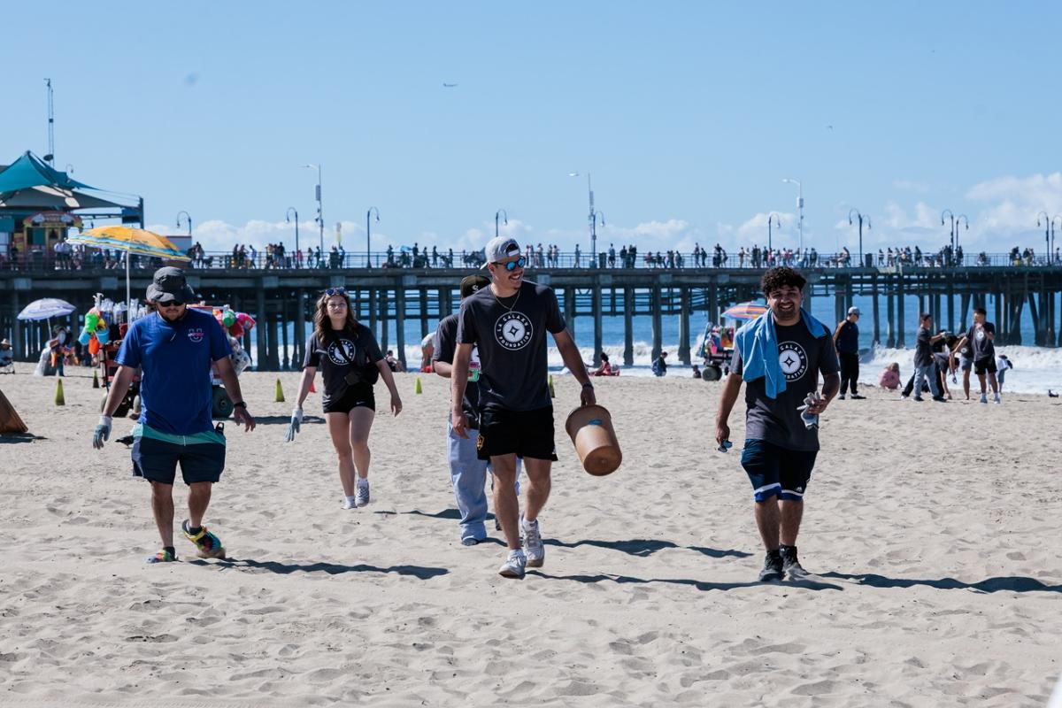 A group of LA Galaxy Foundation volunteers walk across the beach carrying cleanup supplies, with a busy pier and beachgoers behind them.