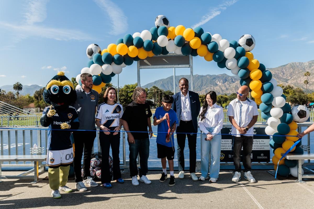 Youth participant cuts a ceremonial ribbon surrounded by LA Galaxy representatives and partners and a team mascot under a balloon arch with mountains in the background.