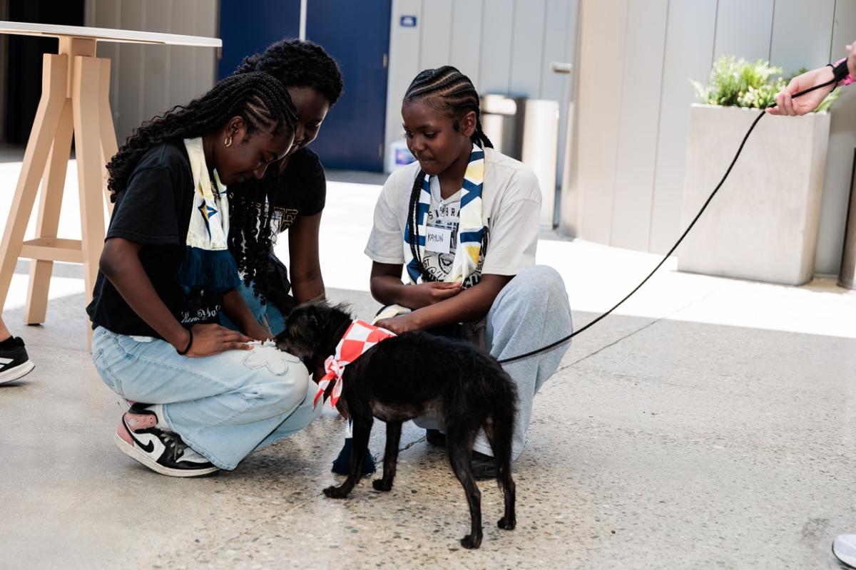 Three young participants sit on the floor petting a small dog wearing a red bandana on a leash.
