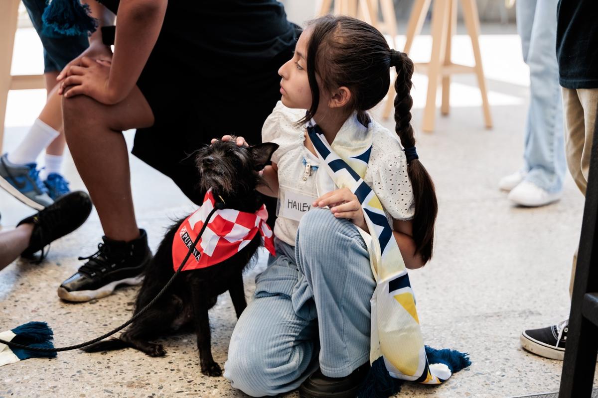 Young participant wearing a scarf sits on the floor next to a small dog in a red bandana, gently petting it.