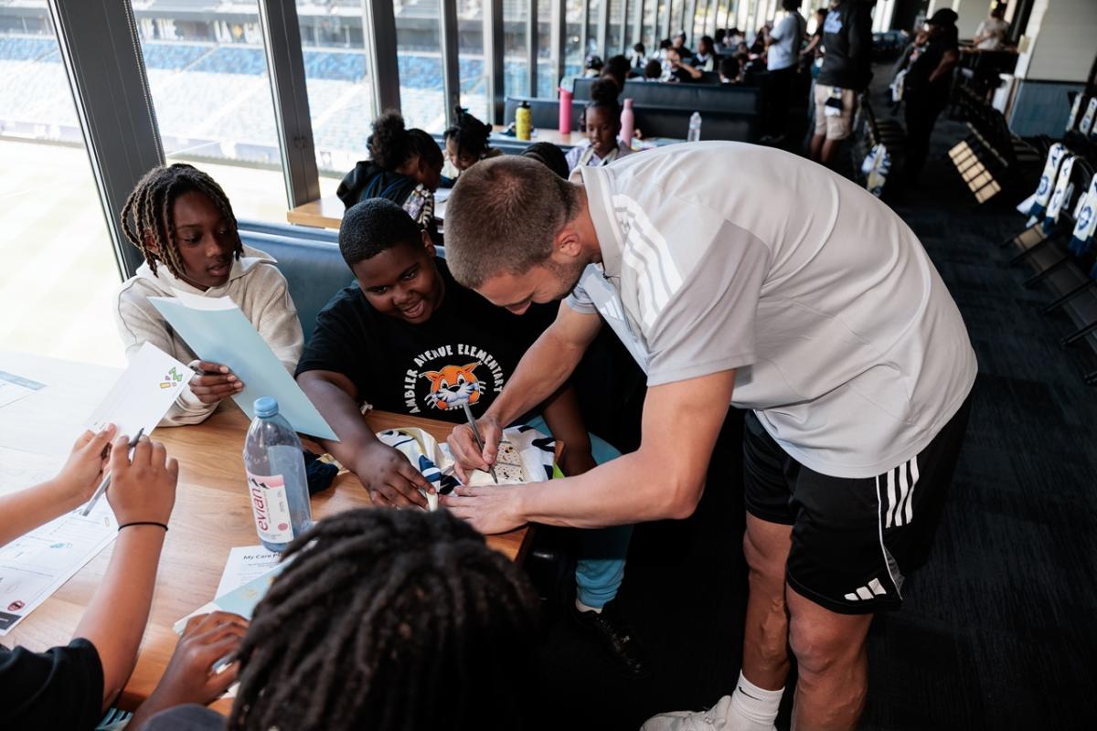 LA Galaxy Defender Chris Rindov signs items while children sit around a table during a hands-on activity session.