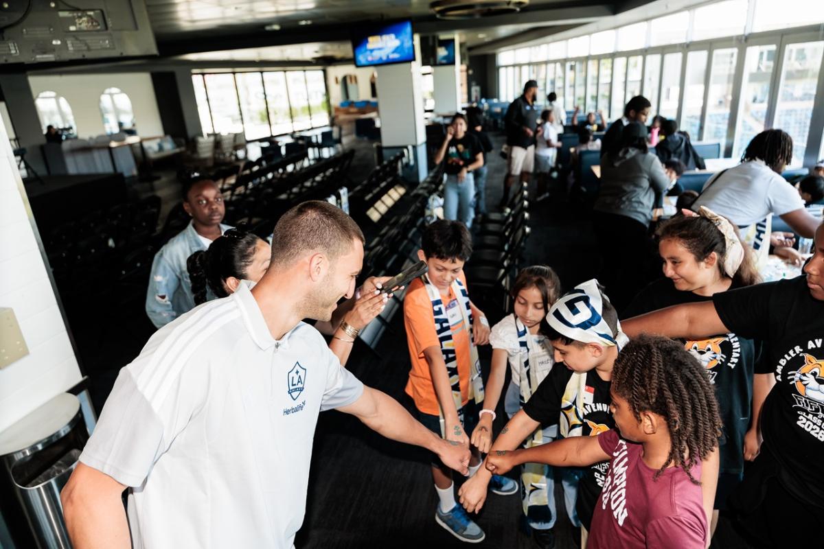 An LA Galaxy Defender Chris Rindov leans in to fist bump a group of kids gathered around a table in a stadium suite.