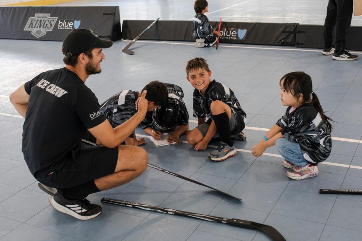 Youth participants in LA Kings ball hockey jerseys gather around a coach during a clinic session on an indoor court. Hockey sticks rest on the floor as participants listen and complete an activity together.