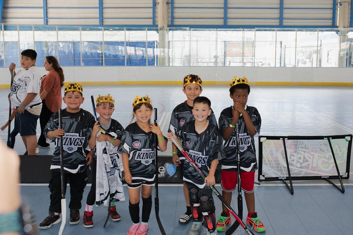 A group of youth participants wearing LA Kings ball hockey jerseys and crowns pose for a team photo on an indoor rink while holding hockey sticks and equipment.