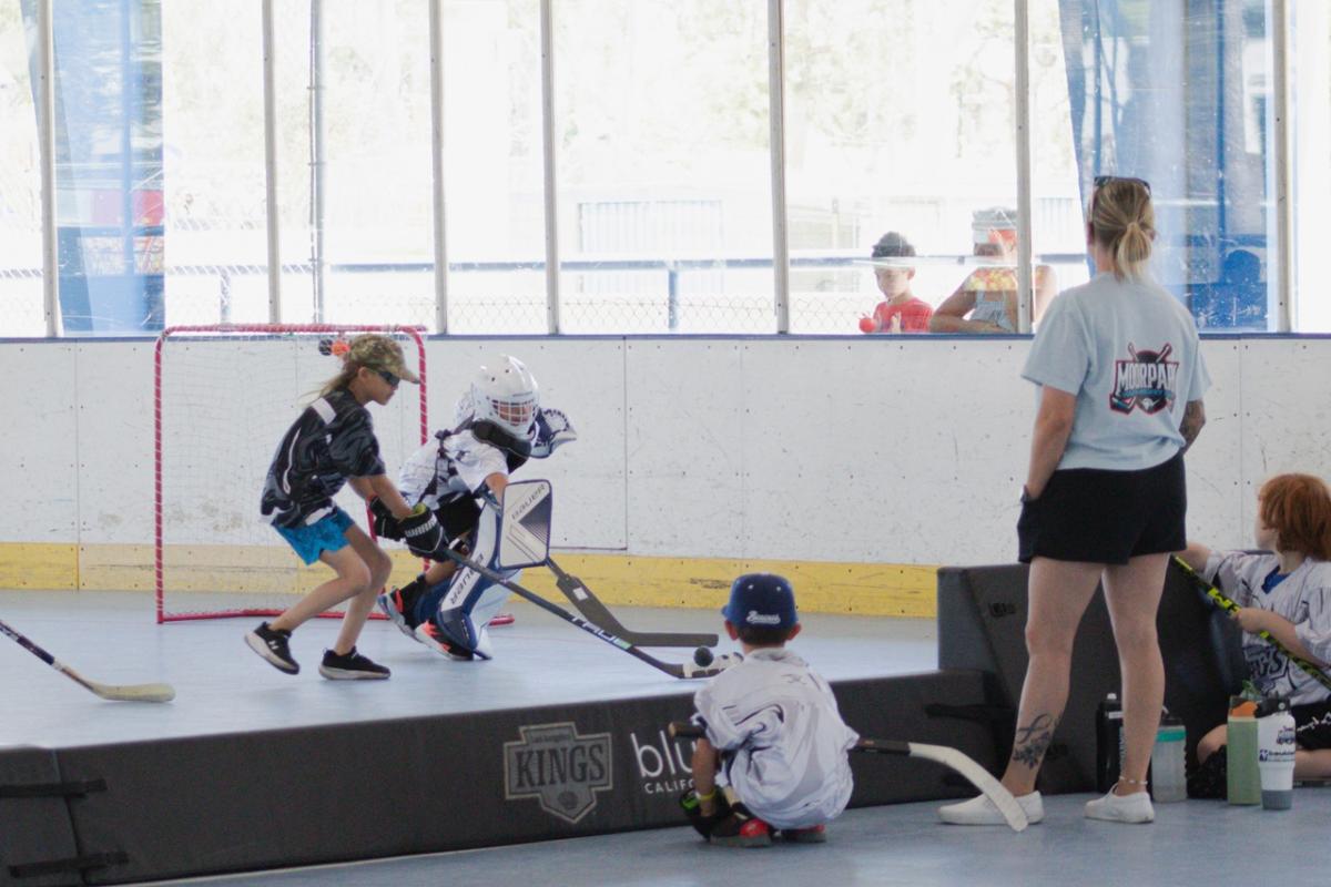 Youth participants compete in a ball hockey game on an indoor rink. One player wearing goalie equipment defends the net while other participants and coaches watch from the sidelines.
