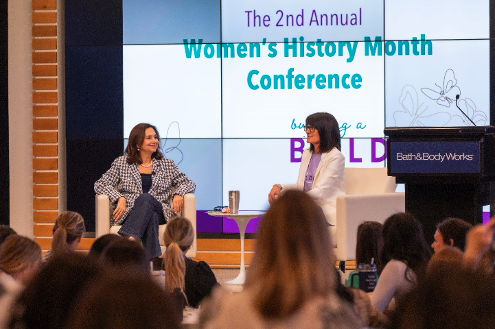 Two women sit together onstage in front of a crowd of employees with a screen behind them.