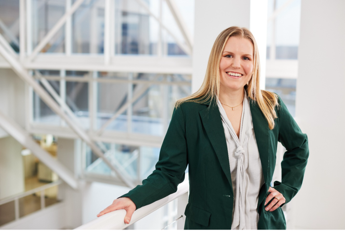 Maureen McHugh smiles at the camera and stands holding a railing at the top of a set of white steps at Bath & Body Works’ home office in Columbus, Ohio. Sun shines through a wall of windows in the background.