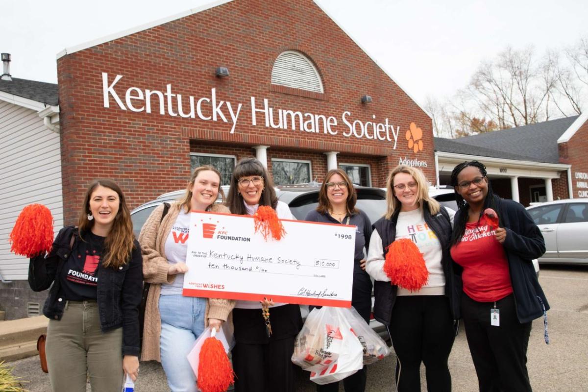 Six people standing together outside in front of the Kentucky Human Society, holding a large check