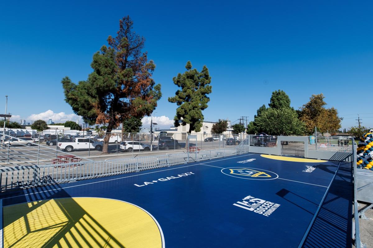 Wide view of a newly installed LA Galaxy-branded mini-pitch with blue and yellow court markings, surrounded by fencing and nearby trees.
