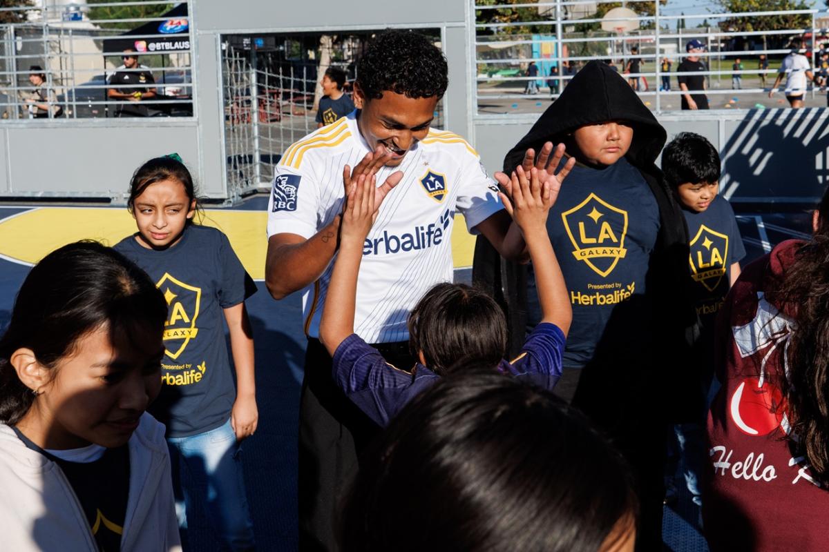 LA Galaxy player in white jersey high-fives youth participants wearing LA Galaxy shirts on a small outdoor soccer court.