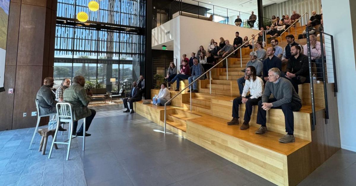 An audience of people sat on a staircase, listening to 3 speakers 