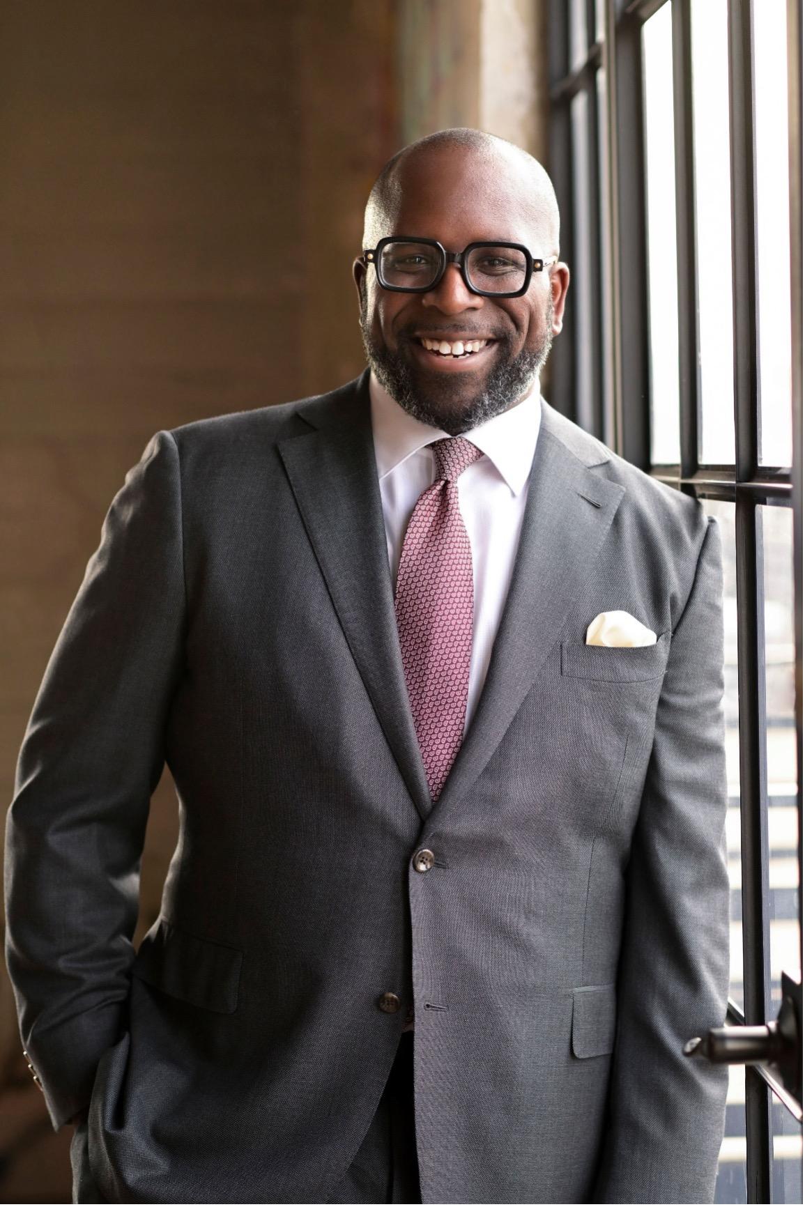 Maurice Cooper is pictured in a grey suit leaning against a sunny window and smiling directly at the camera.