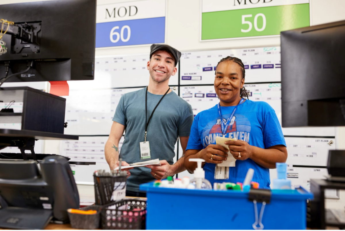Two Bath & Body Works associates stand smiling behind a table with computer monitors and office supplies at a Bath & Body Works distribution center in Columbus, Ohio. A large calendar is posted behind them.