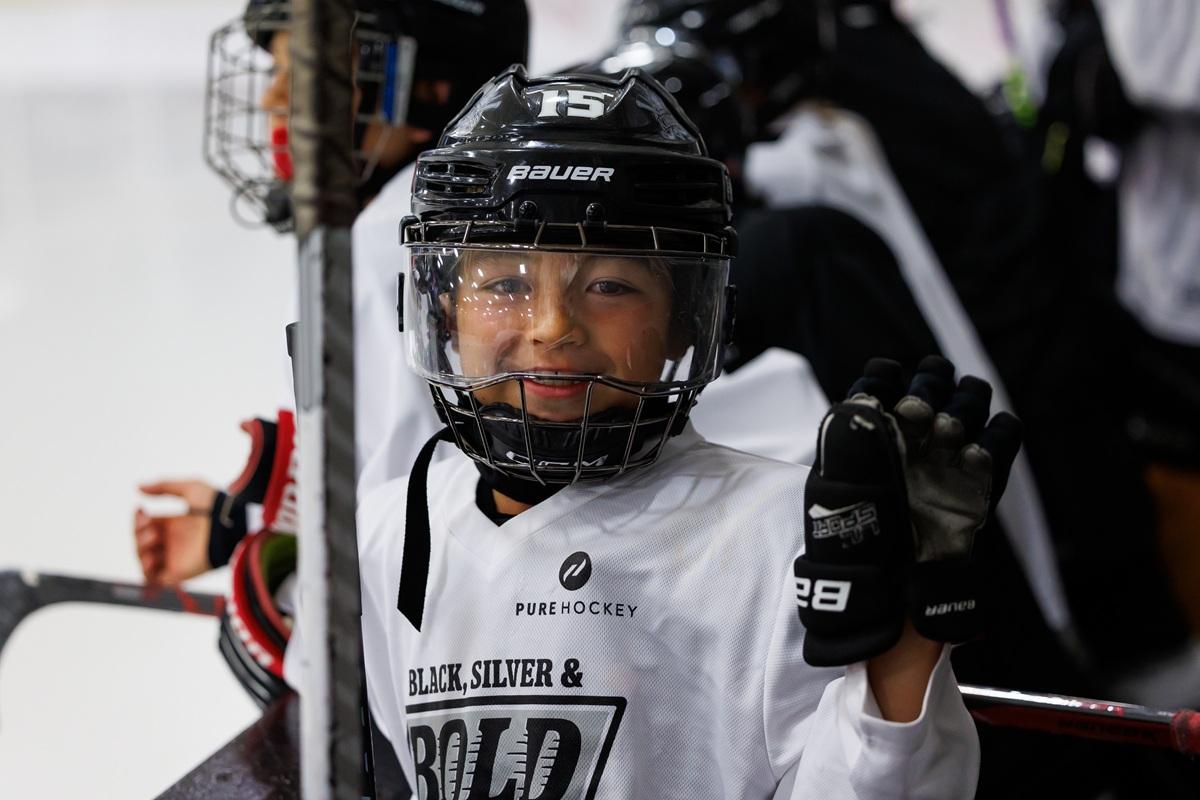 Close-up of a young hockey participant in a white Black, Silver & Bold jersey smiling and waving from the bench.