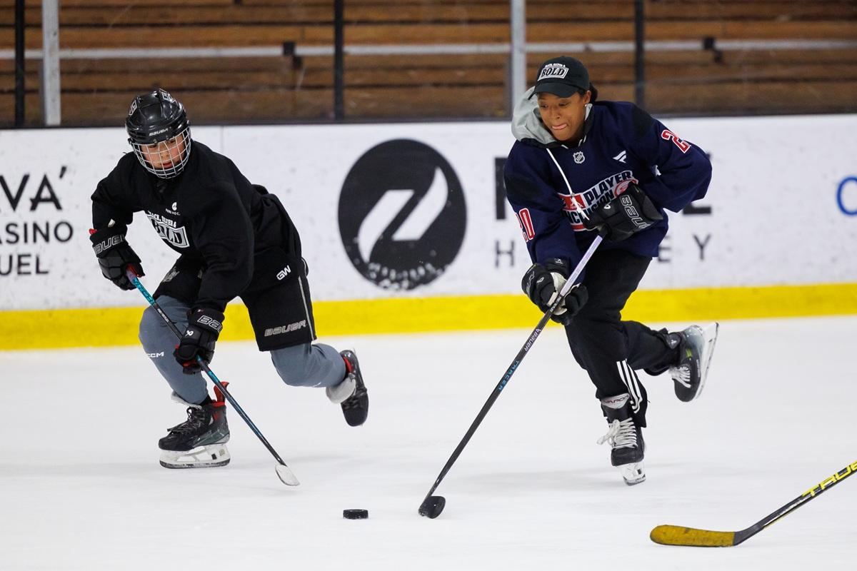 One youth players race for the puck during a Black, Silver & Bold hockey session while a Blake Bolden skates alongside.