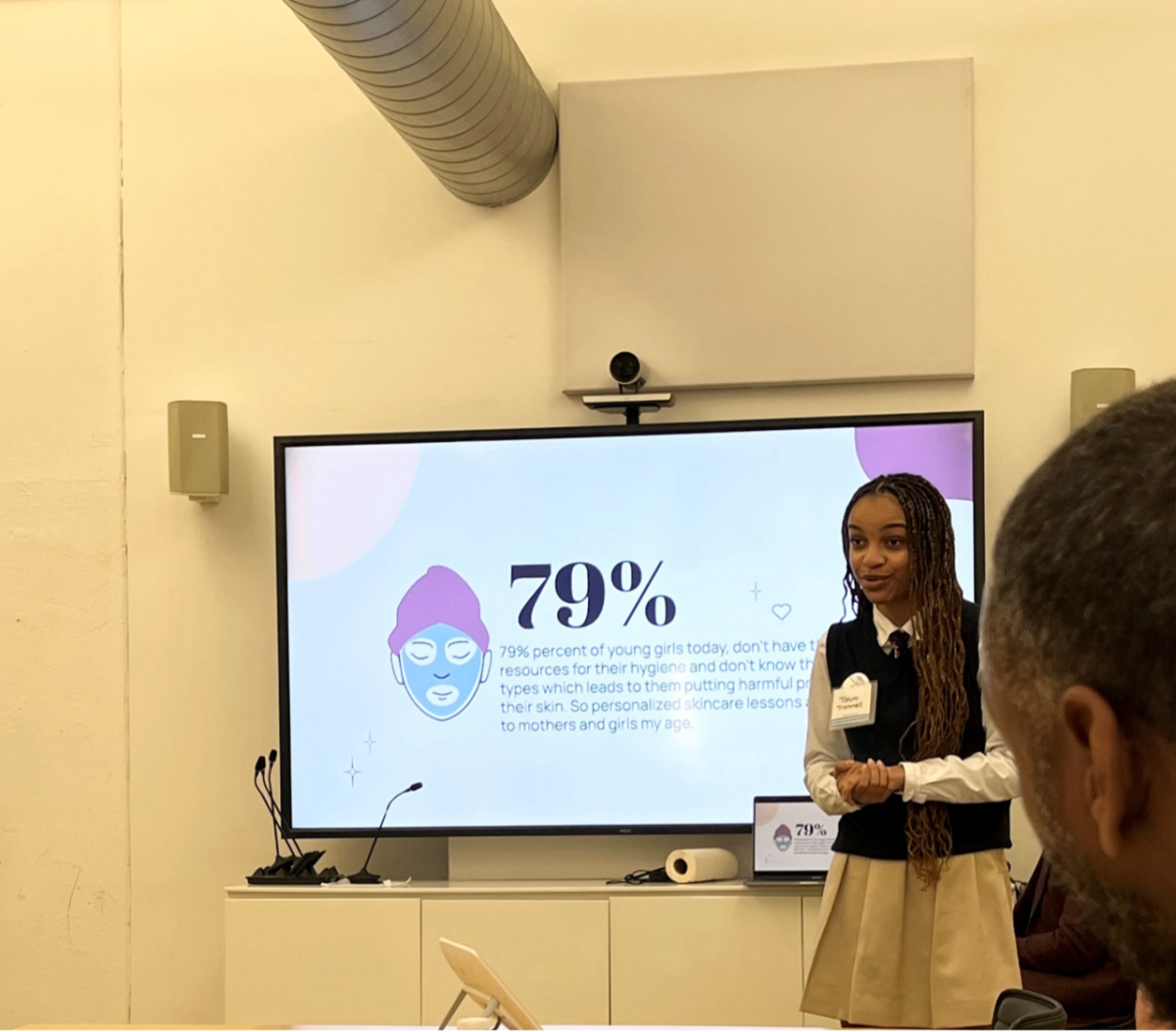 A female middle school student stands in front of a screen and presents information to a group. The screen shows an illustrated woman with her hair wrapped in a towel and a face mask.