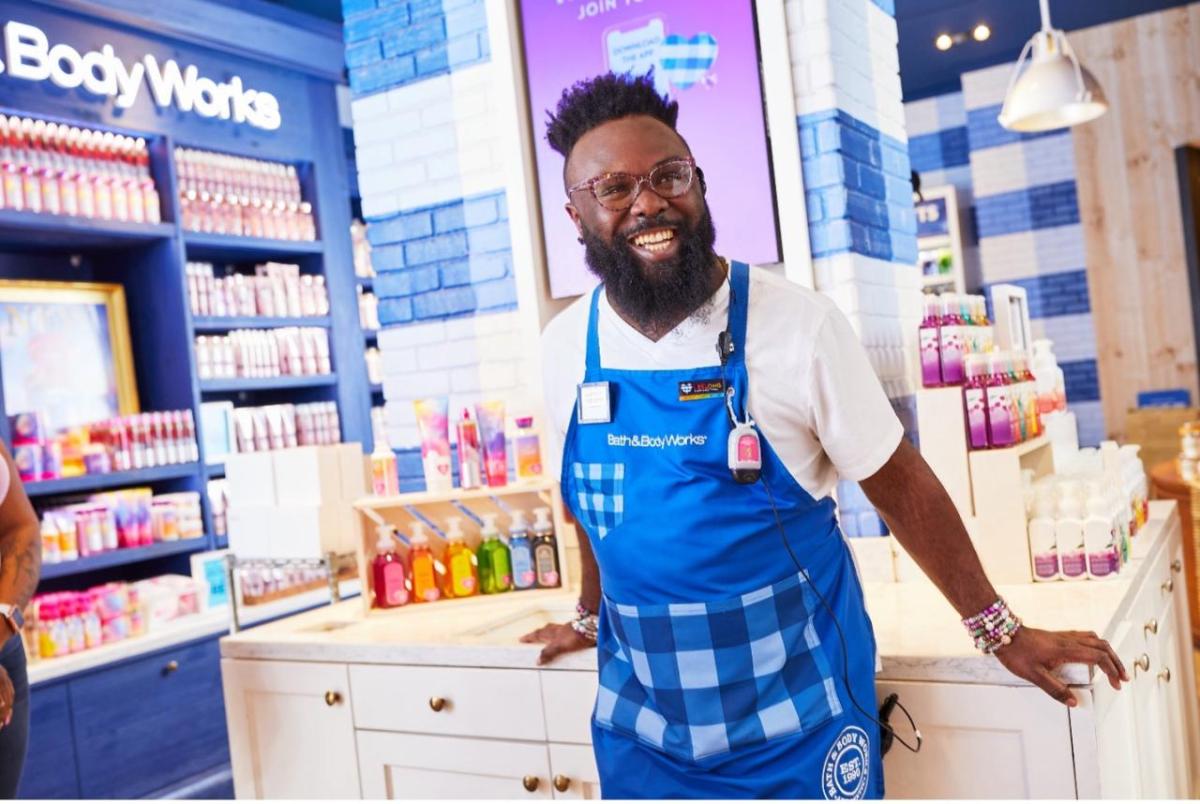 A male Bath & Body Works associate stands in store smiling at the camera in a blue apron with Bath & Body Works products in the background. He leans against a sink where multicolored hand soap bottles are displayed.