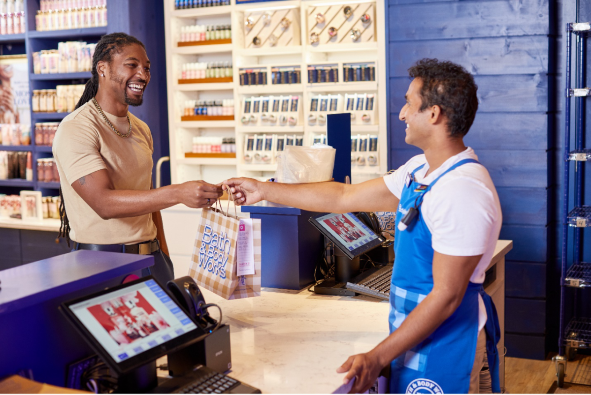A male Bath & Body Works associate stands behind a register in store and hands a smiling customer a Bath & Body Works bag. 