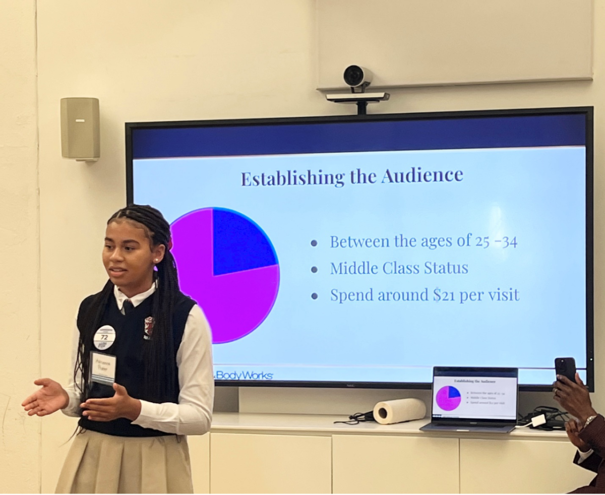 A female middle school student stands in front of a screen and presents information to a group. The screen shows a pie chart with audience data.