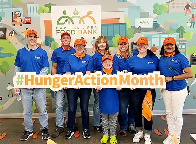 A group of volunteers wearing blue t-shirts and orange caps, holding a 'hunger action month' sign
