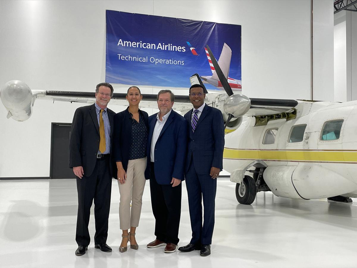Four people standing in front of a small plane in a hanger, American Airlines banner on the wall behind them