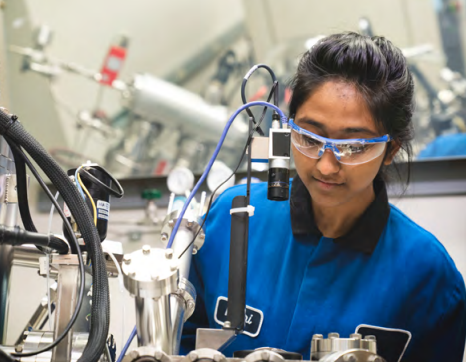 A person in blue and black uniform wearing safety glasses in a lab setting.