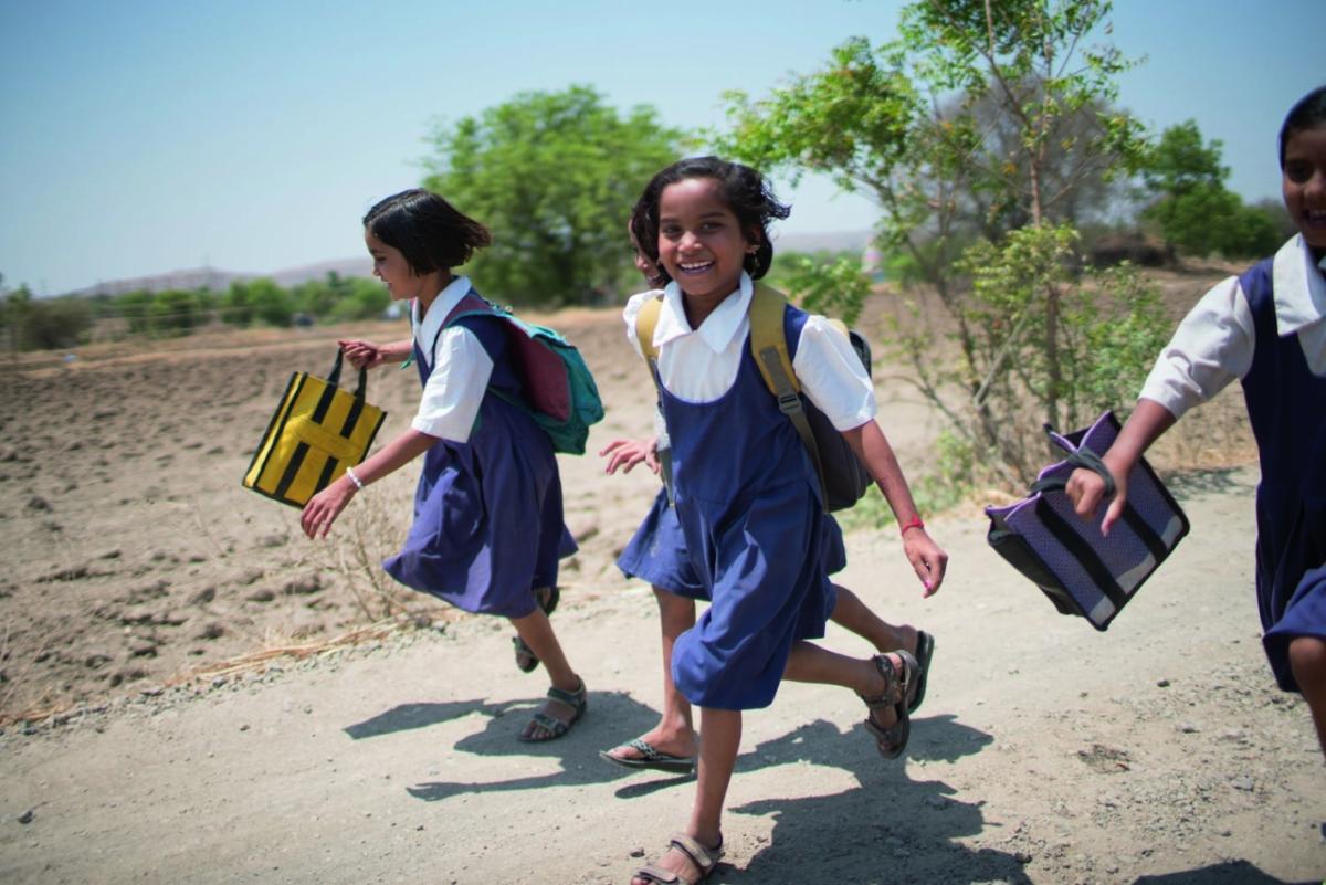 four smiling children running on a dirt road, carrying school bags
