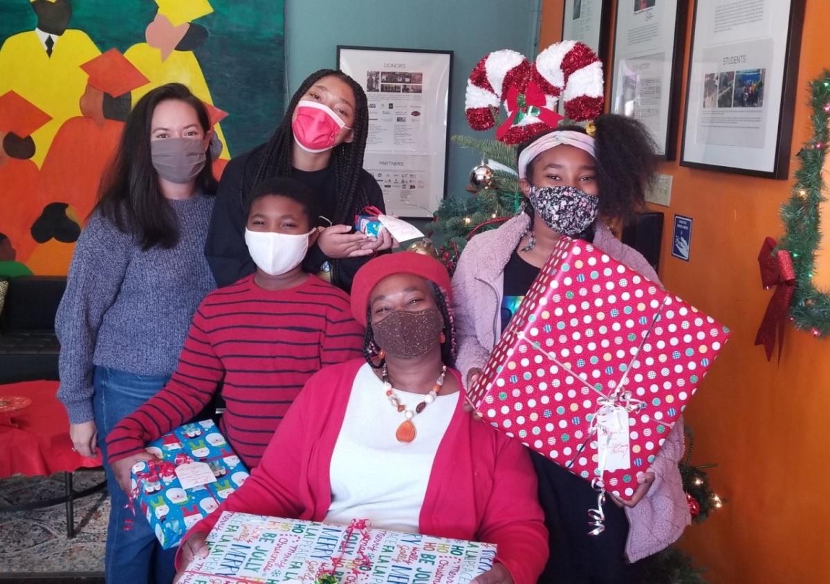 Children holding presents and an AEG employee surround a sitting mother, all wearing masks and smiling. 