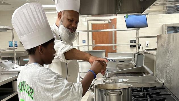 An adult helping a youth cook in a kitchen 