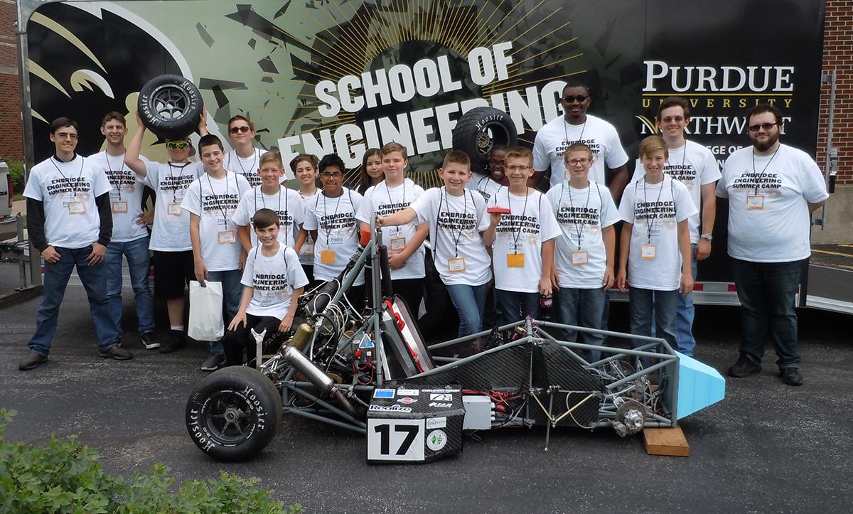 Large group of children stand behind a go-cart type vehicle. All wearing the same School of Engineering t-shirts