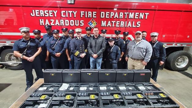 A group of 16 people in uniform stand in front of a Large red vehicle "Jersey City Fire Department Hazardous Materials" on the side. They stand behind 12 cases of equipment.
