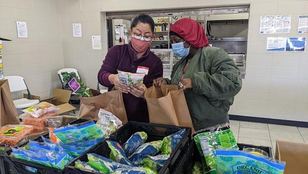 two people look at a bag of vegetables, one holds an open paper bag. They are in a kitchen type setting with crates of bagged vegetables in front of them