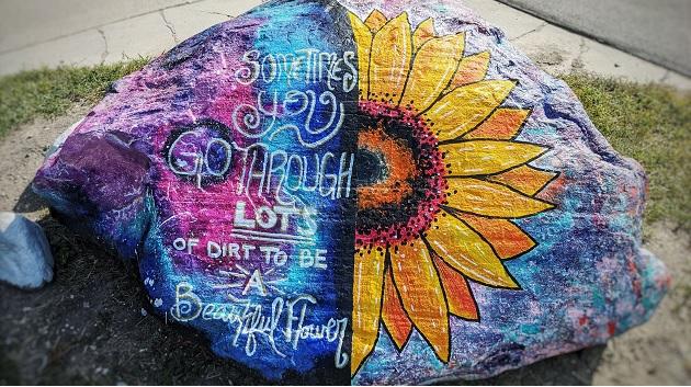 Large painted boulder, right half is half a sunflower the left half is blues and purples with "sometimes you go through lots of dirt to be a beautiful flower"
